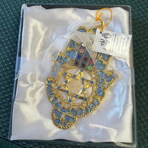 Pode incluir: Um ornamento hamsa de metal dourado com detalhes em esmalte azul, uma estrela central e strass multicoloridos incrustados. Está preso com uma fita branca, inclui uma etiqueta com texto em hebraico e é apresentado numa caixa transparente.