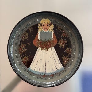 May include: A round ceramic plate featuring a central illustration of a blonde person with curly hair wearing a dark blue apron over a rust-colored long-sleeved shirt and a long white skirt with blue floral accents, surrounded by a dark brown border.