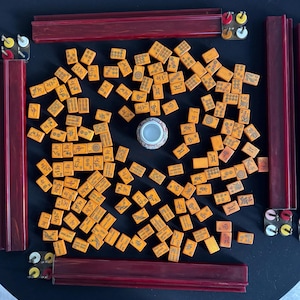May include: A complete Mahjong set with orange tiles featuring Chinese characters and symbols. The set includes four dark red wooden racks, a small decorative container, and tile holders. The tiles are scattered on a black surface, ready for play.