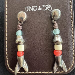 May include: A pair of drop earrings featuring a silver-tone metal stud, a brown cord, a light blue bead, a silver-tone metal bead, a cream bead, a red bead, and a silver-tone metal fish tail charm, displayed on a brown leather card with UNO de 50 text.