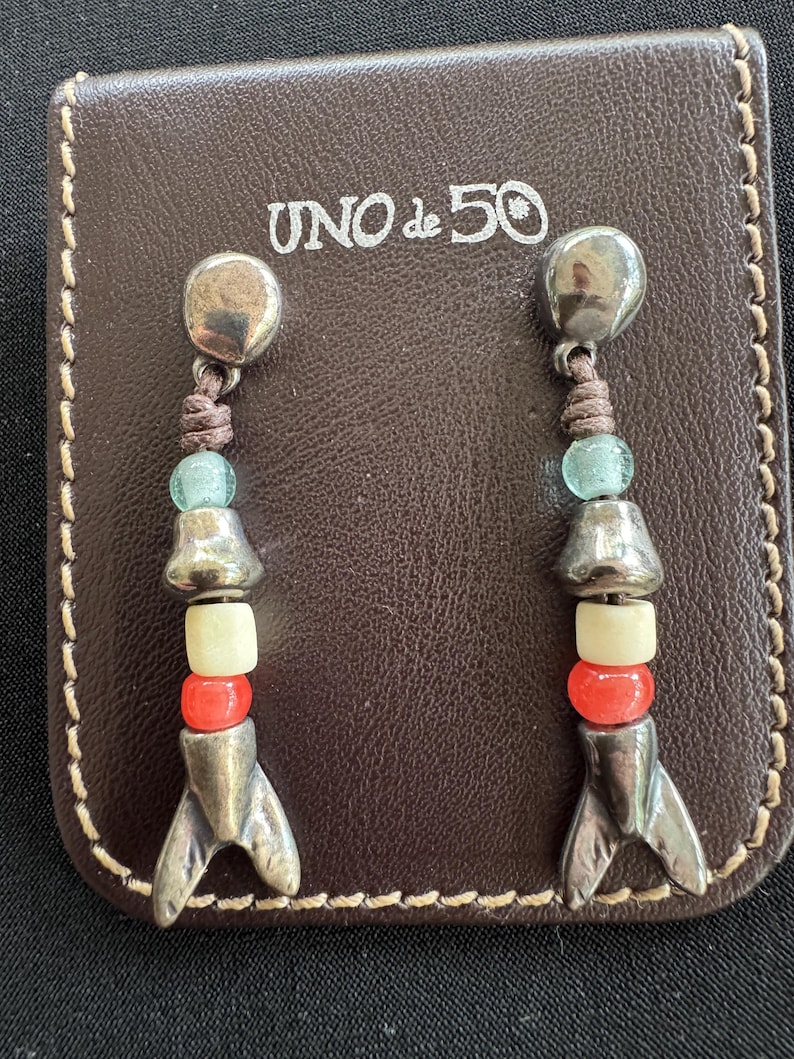 May include: A pair of drop earrings featuring a silver-tone metal stud, a brown cord, a light blue bead, a silver-tone metal bead, a cream bead, a red bead, and a silver-tone metal fish tail charm, displayed on a brown leather card with UNO de 50 text.