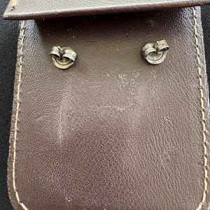 May include: Two silver-toned metal earring backs with a circular design, positioned on a dark brown rectangular leather pouch featuring visible light-colored stitching along the edges and a folded flap at the top of the accessory holder.
