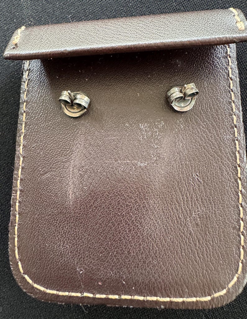 May include: Two silver-toned metal earring backs with a circular design, positioned on a dark brown rectangular leather pouch featuring visible light-colored stitching along the edges and a folded flap at the top of the accessory holder.