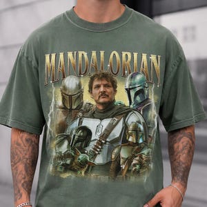 May include: Moss green t-shirt with a graphic of characters from the Mandalorian series. The word "MANDALORIAN" is printed across the top in gold lettering. The shirt has a vintage style and a distressed appearance.