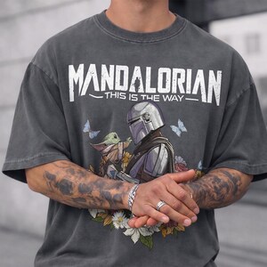 May include: Charcoal grey t-shirt with the white text "MANDALORIAN THIS IS THE WAY". The design shows the Mandalorian and Grogu with flowers and butterflies. The word "PEPPER" is printed at the bottom of the shirt.