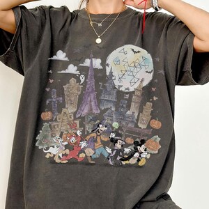 May include: Dark grey t-shirt featuring a Halloween-themed graphic with cartoon characters, a full moon, and spooky buildings. The word "PEPPER" is printed at the bottom of the shirt.