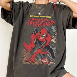 May include: Dark gray t-shirt featuring a comic book graphic of Spider-Man. The design includes the text "BRAND NEW DAY the AMAZING SPIDER-MAN" in red and yellow. The shirt has a vintage, worn look.