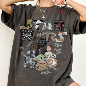 Könnte beinhalten: Dunkelgraues Star Wars Grafik-T-Shirt mit Figuren wie Darth Vader, Baby Yoda und Prinzessin Leia. Das Star Wars-Logo befindet sich oben. Das Wort "Pepper" steht unten.