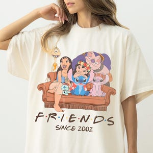 May include: Ivory-coloured t-shirt with a graphic of Lilo & Stitch characters on a brown sofa. The word "FRIENDS" is printed above "SINCE 2002" in black. The shirt has a loose fit. The word "IVORY" is printed at the bottom.