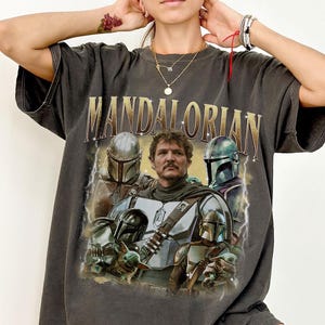May include: Dark grey t-shirt with a vintage graphic of characters from the Mandalorian series. The word "MANDALORIAN" is printed across the top in gold lettering. The shirt has a distressed appearance.