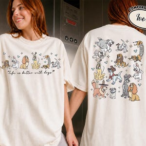 Puede incluir: Camiseta color marfil con un dise&ntilde;o de perros. La parte delantera presenta perros de dibujos animados y el texto "Life is better with dogs." La parte trasera muestra varias ilustraciones de perros. La camiseta es de la marca Comfort Colors.