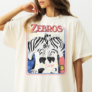 Op de afbeelding: Ivoor kleurig T-shirt met een afbeelding van twee zebra's met zwart-witte strepen, omlijst in een rode en blauwe rand. Het woord "ZEBROS" staat in een retro lettertype boven de zebra's gedrukt. Het shirt heeft een relaxte pasvorm.