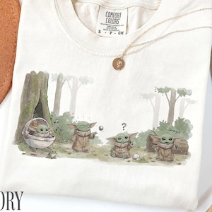 Pu&ograve; includere: T-shirt color avorio con grafica di Grogu. Il design mostra Grogu in un ambiente forestale. La maglietta ha girocollo e maniche corte. Una collana d'oro con un ciondolo &egrave; drappeggiata sulla maglietta.