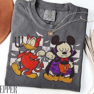 以下が含まれることがあります： チャコールグレーのクルーネックTシャツ。赤い悪魔の仮装をしたアヒルのキャラクターと、吸血鬼の仮装をしたネズミのキャラクターのグラフィックがプリントされています。首元にはComfort Colorsのタグがあり、細いゴールドのネックレスが上に置かれています。