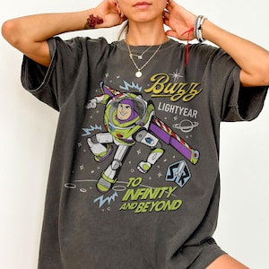 Pode incluir: T-shirt cinzenta escura com um desenho de Buzz Lightyear e as frases "Buzz Lightyear" e "To Infinity and Beyond". O design inclui um tema espacial com estrelas e planetas. A t-shirt tem um corte folgado.