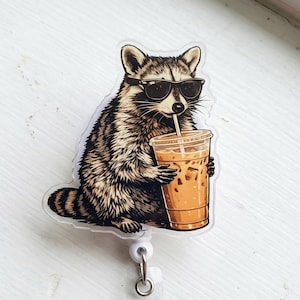 May include: A badge reel featuring a raccoon wearing sunglasses, holding a cup of iced coffee with a straw. The raccoon is brown, black, and white. The cup is clear with a brown beverage and ice cubes. The badge reel has a clear plastic base.