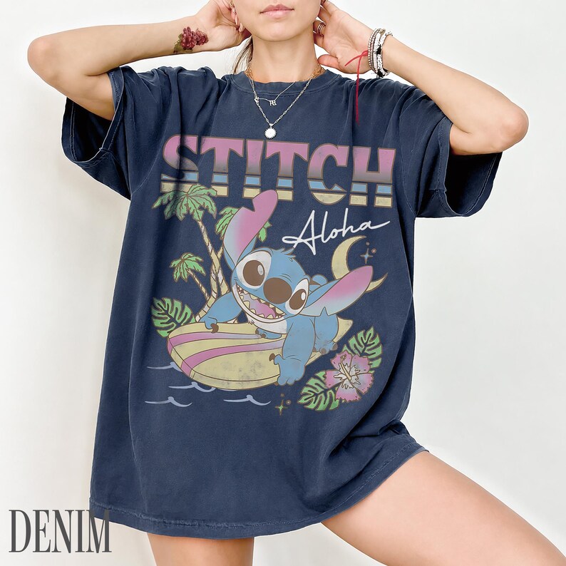 May include: Navy blue t-shirt featuring a graphic of Stitch surfing with the text "STITCH Aloha" in a retro style. The design includes palm trees, a crescent moon, and tropical flowers. The word "DENIM" is printed at the bottom.