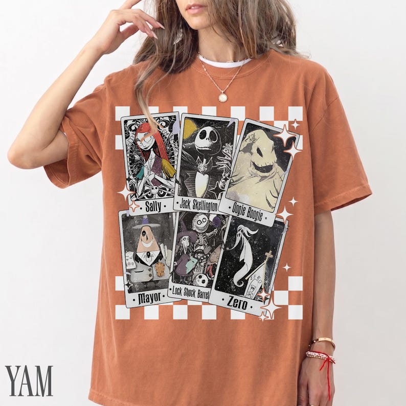 May include: Rust-colored t-shirt featuring a graphic of playing cards with characters from The Nightmare Before Christmas. The cards include Sally, Jack Skellington, Oogie Boogie, Mayor, Lock Shock Barrel, and Zero. The shirt has a white checkered pattern.
