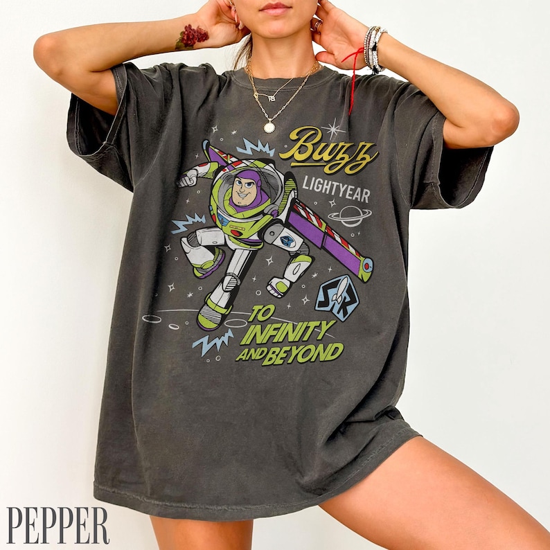 Pode incluir: T-shirt cinzenta escura com um desenho de Buzz Lightyear e as frases "Buzz Lightyear" e "To Infinity and Beyond". O design inclui um tema espacial com estrelas e planetas. A t-shirt tem um corte folgado.