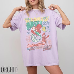 Puede incluir: Una camiseta de manga corta de color morado claro con un estampado gr&aacute;fico de Ariel, Flounder y Sebasti&aacute;n. El texto dice Atlantica Ocean Tour 1991, Sebastian with the Hot Crustacean Band. El dise&ntilde;o incluye ilustraciones de coral y burbujas.