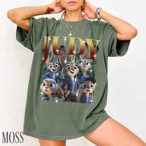May include: Green t-shirt featuring a graphic of Judy Hopps from Zootopia. The design includes the name "JUDY" in gold lettering with a red outline, and the word "Hopps" in a script font. The shirt has a vintage, washed look.