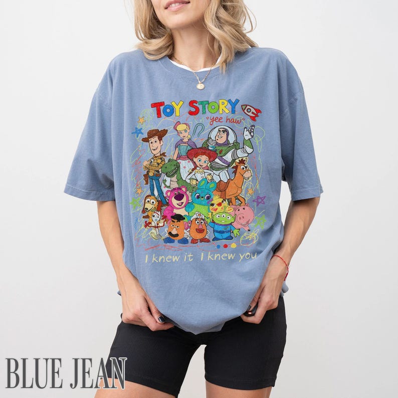 May include: A dusty blue short-sleeve t-shirt featuring a colourful graphic print of Toy Story characters including Woody, Buzz Lightyear, and Jessie. The text reads Toy Story, yee haw, and I knew it I knew you, surrounded by small star and rocket doodles.