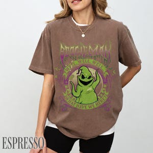 May include: Brown t-shirt with a green cartoon character design and the words "BOOGIEMAN", "WELL, WELL, WELL... WHAT HAVE WE HERE?" in green and purple. The shirt is a dark brown colour.
