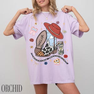 May include: A light purple t-shirt featuring a graphic print of a cowboy boot with a cow print pattern, a red hat, and blocks labeled with the letters T, S, and the number 13. The design includes the name Taylor, a beaded string, and flowers.