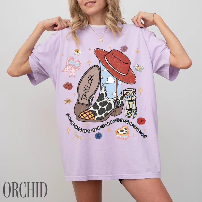 May include: A light purple t-shirt featuring a graphic print of a cowboy boot with a cow print pattern, a red hat, and blocks labeled with the letters T, S, and the number 13. The design includes the name Taylor, a beaded string, and flowers.
