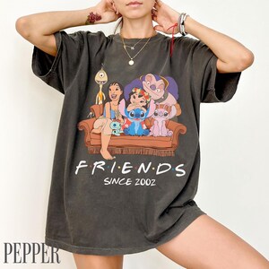 May include: Dark grey t-shirt with a cartoon graphic of Lilo & Stitch characters on a brown sofa. The word "FRIENDS" is printed in white, with "SINCE 2002" underneath. The t-shirt is a casual wear item.