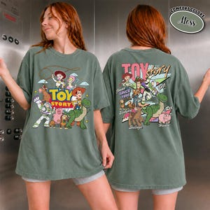 K&ouml;nnte beinhalten: Moosgr&uuml;nes T-Shirt mit einem Toy Story-Motiv. Es zeigt Woody, Buzz Lightyear und Jessie. Auf der Vorderseite ist das Toy Story-Logo zu sehen. Auf der R&uuml;ckseite sind weitere Charaktere und der Text "Jessie Since 1995".