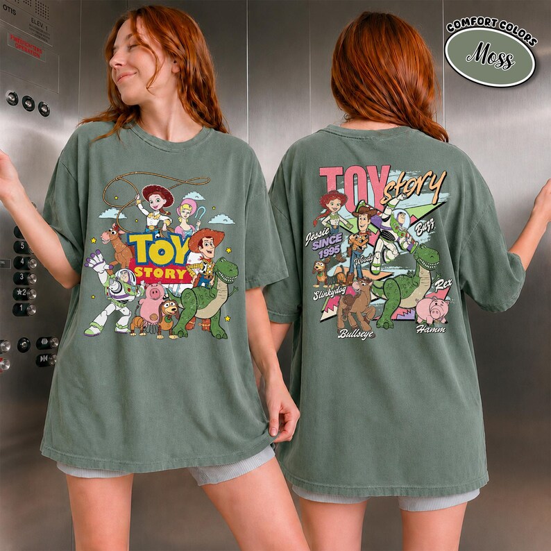 K&ouml;nnte beinhalten: Moosgr&uuml;nes T-Shirt mit einem Toy Story-Motiv. Es zeigt Woody, Buzz Lightyear und Jessie. Auf der Vorderseite ist das Toy Story-Logo zu sehen. Auf der R&uuml;ckseite sind weitere Charaktere und der Text "Jessie Since 1995".