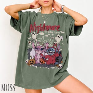 May include: A moss green short-sleeve t-shirt featuring a graphic print with the text Nightmare on Main Street. The design includes two cartoon characters, one pink and one blue, dressed in Halloween costumes, surrounded by ghosts, pumpkins, and a spiderweb.