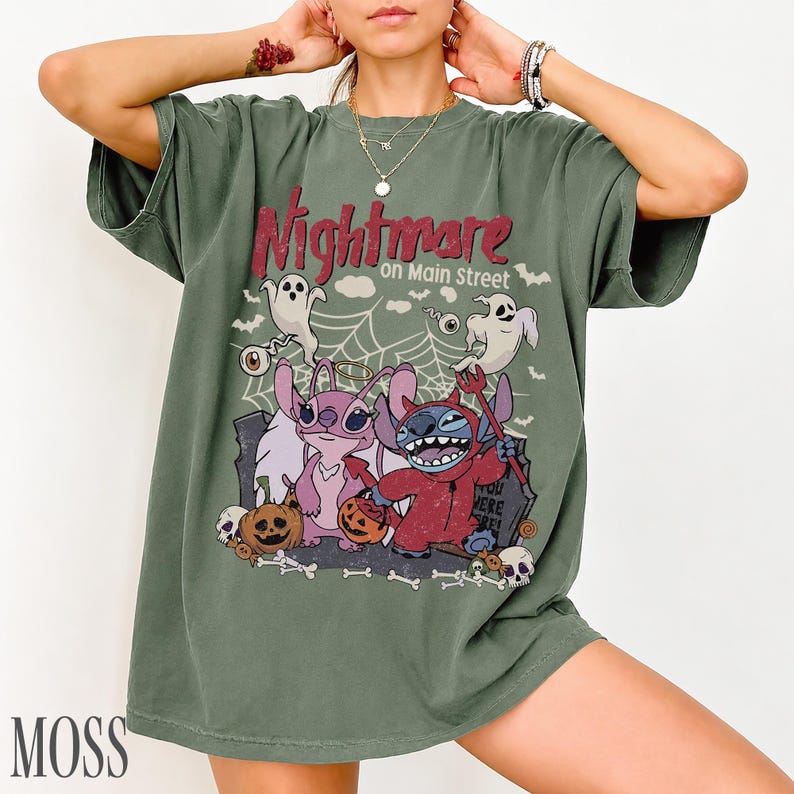 May include: A moss green short-sleeve t-shirt featuring a graphic print with the text Nightmare on Main Street. The design includes two cartoon characters, one pink and one blue, dressed in Halloween costumes, surrounded by ghosts, pumpkins, and a spiderweb.