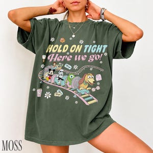 May include: Moss green t-shirt with a colorful graphic of a train carrying cartoon characters. The text "HOLD ON TIGHT Here we go!" is printed above the train. White flower and star accents surround the graphic.