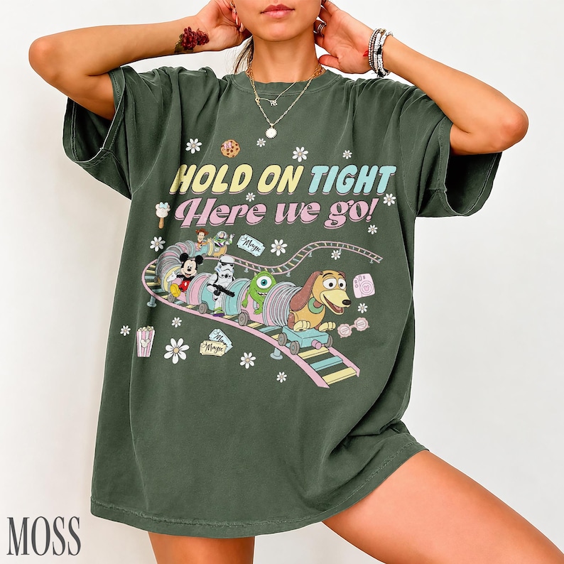 May include: Moss green t-shirt with a colorful graphic of a train carrying cartoon characters. The text "HOLD ON TIGHT Here we go!" is printed above the train. White flower and star accents surround the graphic.