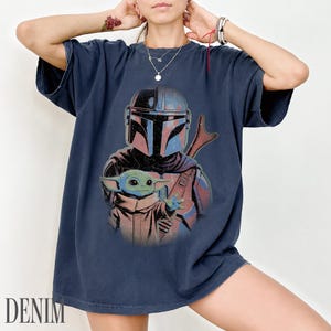 Puede incluir: Una camiseta azul marino de manga corta con un estampado gr&aacute;fico de un personaje mandaloriano con casco y una peque&ntilde;a criatura verde de orejas grandes. El dise&ntilde;o tiene un aspecto desgastado con acentos de color rojo y azul apagados.
