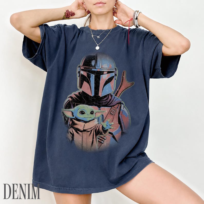 Puede incluir: Una camiseta azul marino de manga corta con un estampado gr&aacute;fico de un personaje mandaloriano con casco y una peque&ntilde;a criatura verde de orejas grandes. El dise&ntilde;o tiene un aspecto desgastado con acentos de color rojo y azul apagados.
