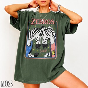 Op de afbeelding: Mosgroen T-shirt met een retro afbeelding van twee zebra's en de tekst "Zebros". De zebra's zijn zwart en wit, met blauwe, roze en gele accenten. Het shirt heeft een relaxte pasvorm.
