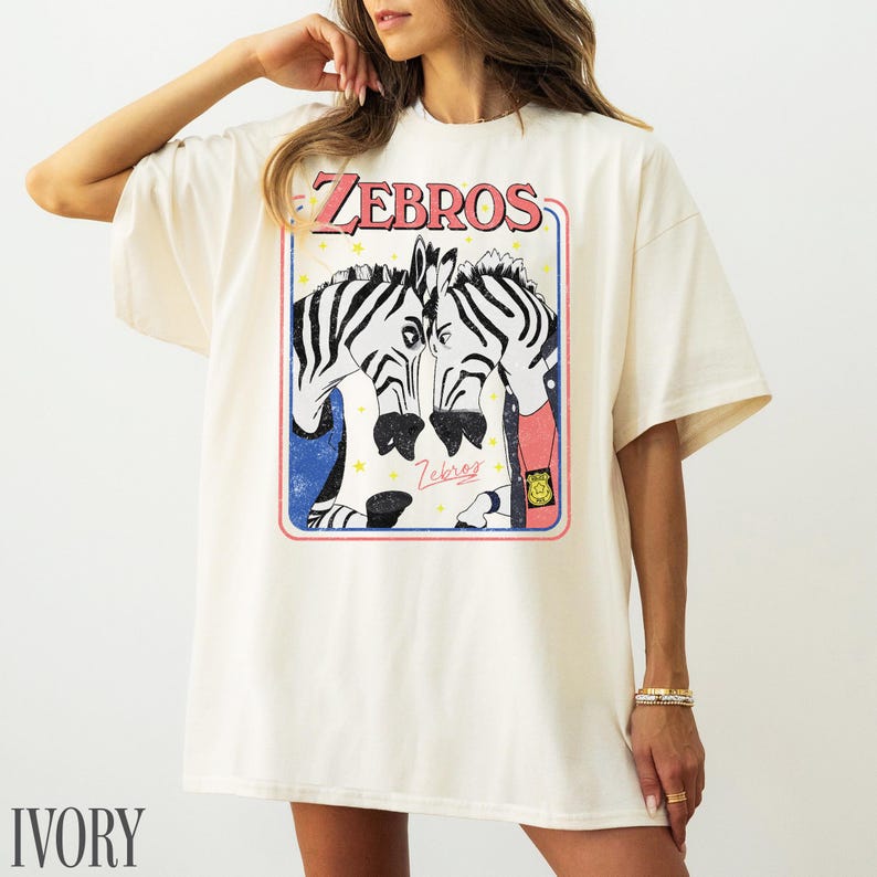 Op de afbeelding: Ivoor kleurig T-shirt met een afbeelding van twee zebra's met zwart-witte strepen, omlijst in een rode en blauwe rand. Het woord "ZEBROS" staat in een retro lettertype boven de zebra's gedrukt. Het shirt heeft een relaxte pasvorm.