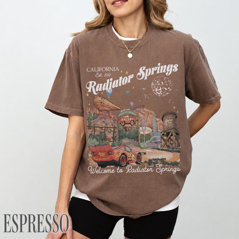 May include: A brown short-sleeve t-shirt featuring a graphic print of Radiator Springs with a red race car, desert rock formations, and a water tower. The text reads California Est. 2012, Radiator Springs, and Welcome to Radiator Springs.