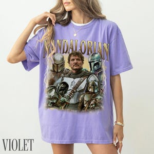 May include: Violet t-shirt with a graphic of characters from The Mandalorian. The design includes the word "MANDALORIAN" in gold lettering above a portrait of the characters. The shirt is a comfortable, casual style.