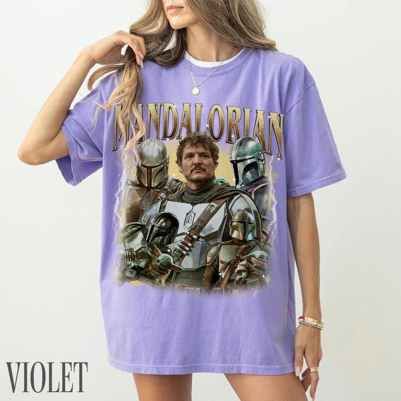 May include: Violet t-shirt with a graphic of characters from The Mandalorian. The design includes the word "MANDALORIAN" in gold lettering above a portrait of the characters. The shirt is a comfortable, casual style.
