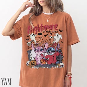 May include: A burnt orange short-sleeve t-shirt featuring a graphic print with the text "Nightmare on Main Street". The design includes two cartoon characters, ghosts, spiderwebs, pumpkins, and skulls in a Halloween-themed illustration.