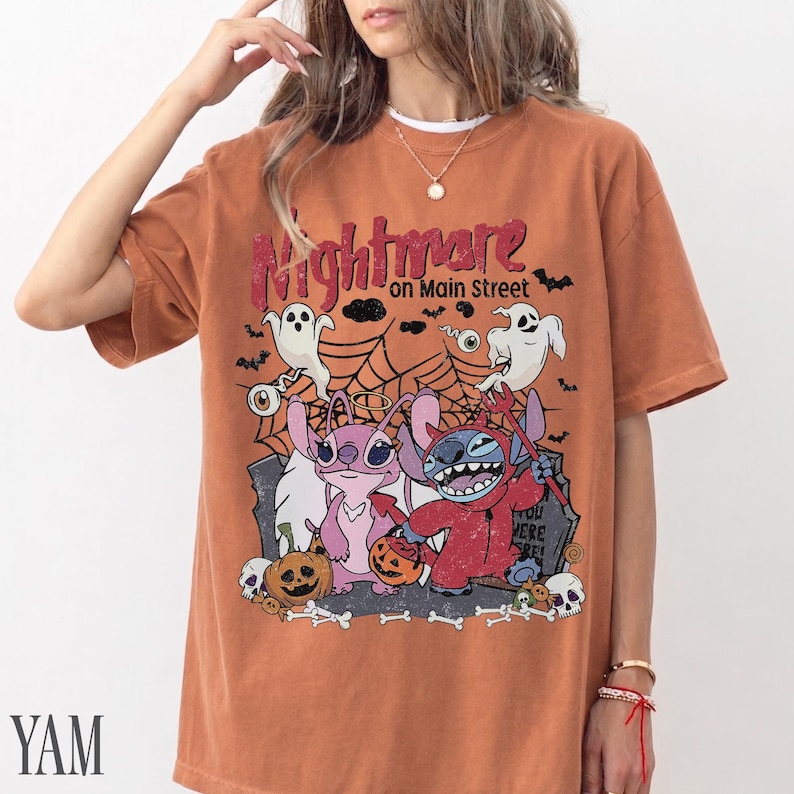 May include: A burnt orange short-sleeve t-shirt featuring a graphic print with the text "Nightmare on Main Street". The design includes two cartoon characters, ghosts, spiderwebs, pumpkins, and skulls in a Halloween-themed illustration.