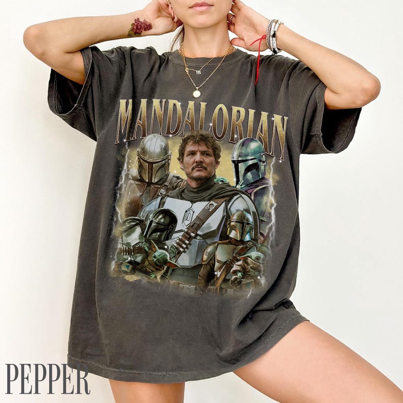 May include: Dark grey t-shirt with a vintage graphic of characters from the Mandalorian series. The word "MANDALORIAN" is printed across the top in gold lettering. The shirt has a distressed appearance.