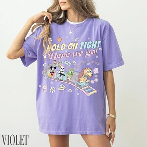 May include: Violet-colored t-shirt with the text "HOLD ON TIGHT Here we go!" and a cartoon graphic of Disney characters on a roller coaster. The graphic includes Mickey Mouse, a Stormtrooper, and Slinky Dog.
