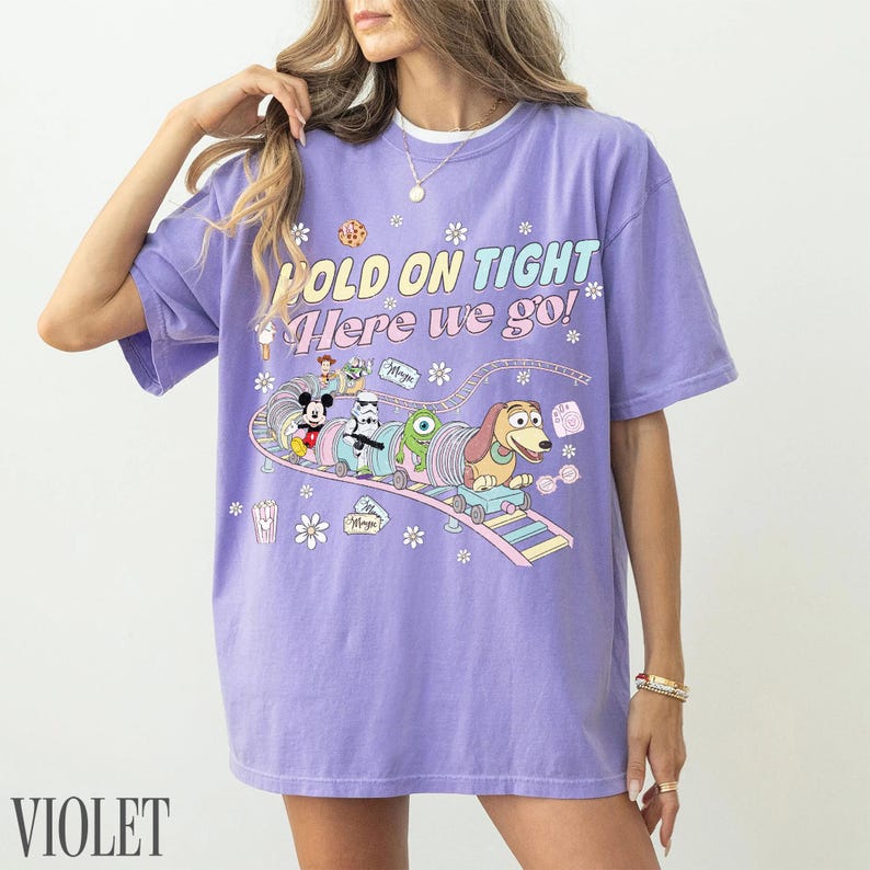 May include: Violet-colored t-shirt with the text "HOLD ON TIGHT Here we go!" and a cartoon graphic of Disney characters on a roller coaster. The graphic includes Mickey Mouse, a Stormtrooper, and Slinky Dog.