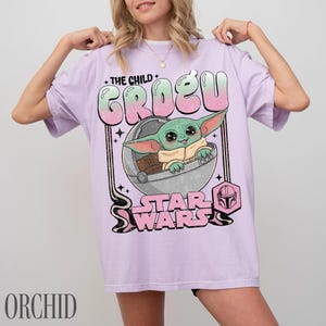 May include: A light purple t-shirt featuring a graphic print of the character Grogu from Star Wars sitting in a floating pod. The text reads THE CHILD GROGU and STAR WARS in a stylized font with a pink and green gradient. The design includes small stars.