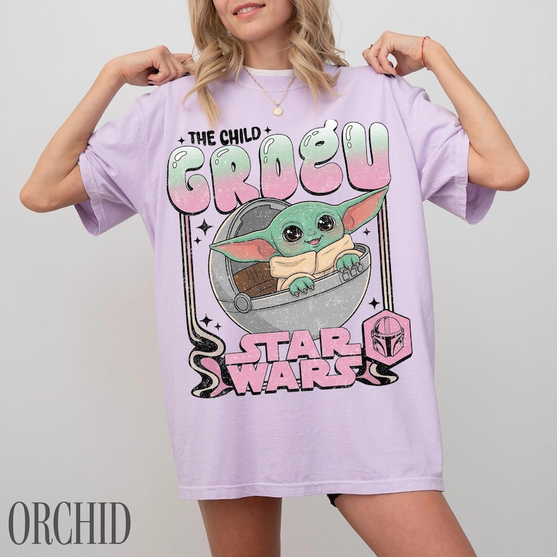 May include: A light purple t-shirt featuring a graphic print of the character Grogu from Star Wars sitting in a floating pod. The text reads THE CHILD GROGU and STAR WARS in a stylized font with a pink and green gradient. The design includes small stars.